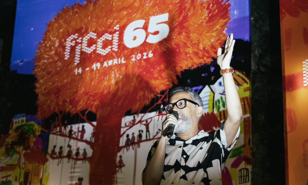 Kike Sierra sostiene un micrófono y levanta una mano durante la presentación oficial del afiche del FICCI 65, frente a una gran imagen colorida del árbol que protagoniza el cartel, con las fechas del festival (14–19 abril 2026) visibles en el fondo.