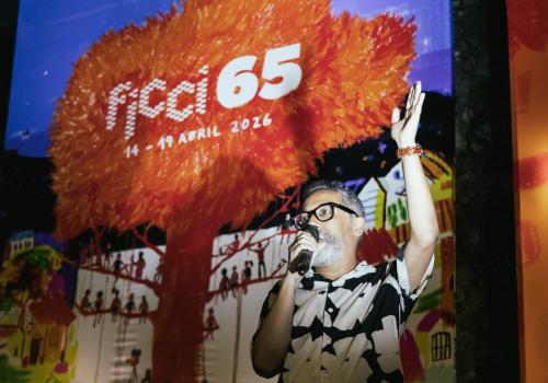 Kike Sierra sostiene un micrófono y levanta una mano durante la presentación oficial del afiche del FICCI 65, frente a una gran imagen colorida del árbol que protagoniza el cartel, con las fechas del festival (14–19 abril 2026) visibles en el fondo.